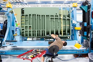 A WORKER LEANS OVER IN AN OFFICE CHAIR TO WORK ON A GIANT PIECE OF MACHINERY