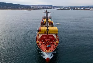 A red container ship stacked with yellow shipping containers, moving through a large expanse of sea with a distant shoreline in the background