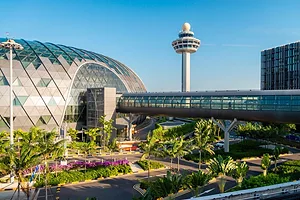 AN OUTSIDE VIEW OF A GLASS AND STEEL AIRPORT TERMINAL, WITH A CONTROL TOWER VISIBLE BEHIND