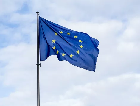 A EUROPEAN UNION FLAG WAVING IN THE WIND ATOP A FLAG POLE IN FRONT OF A CLOUDY, BLUE SKY.