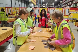 GIRL IN HI VIS VESTS WORK AT A TABLE, OVERSEEN BY AN OLDER INSTRUCTOR