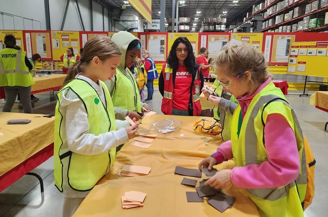 GIRL IN HI VIS VESTS WORK AT A TABLE, OVERSEEN BY AN OLDER INSTRUCTOR