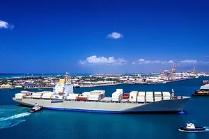 A white ship stacked with white shipping containers moving into a seaside port on a sunny day