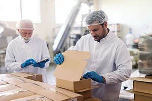 An employee in sterile uniform packing goods in boxes. In background supervisor holding tablet and counting boxes.