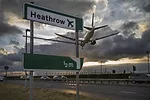 A green sign alongside a road that reads "Heathrow," with a white passenger airplane flying overhead