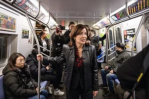 A WOMAN WITH BROWN HAIR, WEARING A LEATHER JACKET, HOLDS ONTO A POLE IN A CROWDED SUBWAY CAR
