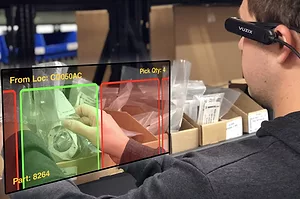 A PERSON FACES A WAREHOUSE PICK WALL USING TECHNOLOGY THAT HIGHLIGHTS THE CORRECT LOCATION IN GREEN