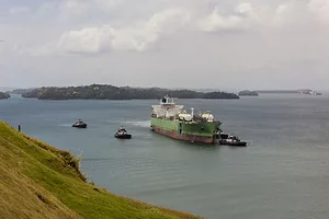 A LARGE GREEN AND WHITE SHIP IS PULLED BY THREE TUGS ACROSS A WATERWAY