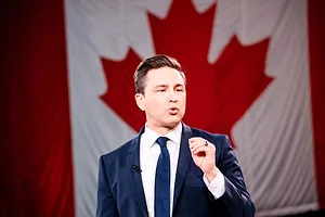A MAN IN A DARK BLUE SUIT AND TIE IN FRONT OF A CANADIAN FLAG PINCHES HIS FINGER AND THUMB TOGETHER IN A GESTURE