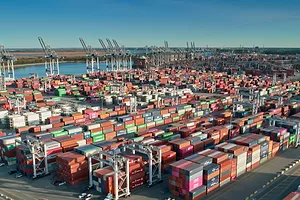 Stacks of multi-colored shipping containers arranged in rows at a port, with rows of blue and grey shipping cranes lining the background along the coastline