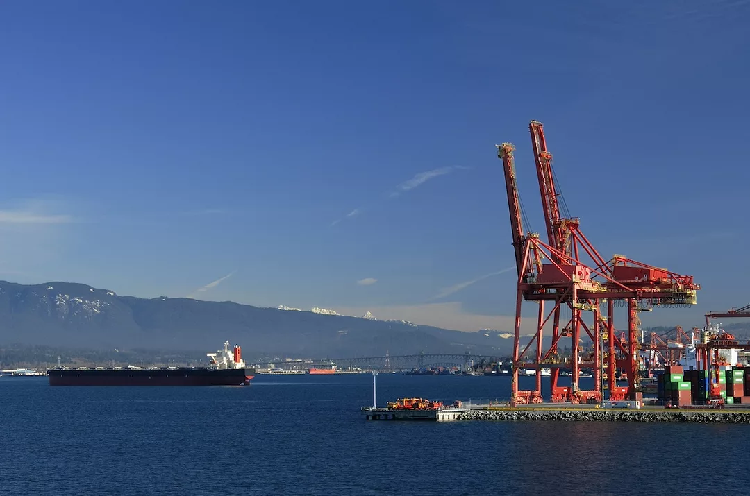 Two large red cranes at a shipping port, with a black container ship moving past the shore.