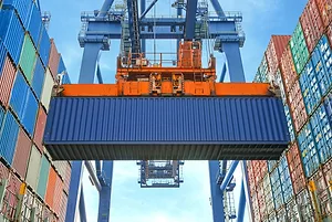 A blue shipping container held by a large blue crane, situated in between stacks of multi-colored shipping containers at a port