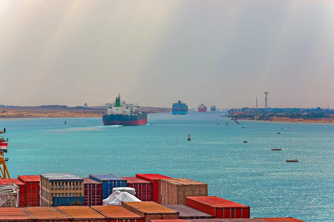 LARGE CONTAINER SHIPS TRAVEL ON THE SUEZ CANAL WITH CONTAINERS IN THE FOREGROUND OF THE PHOTO.