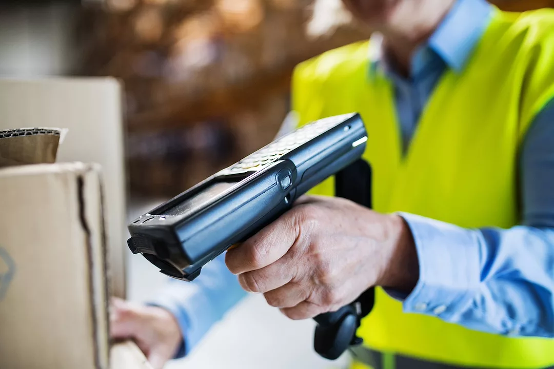 A man in a yellow vest with a black handheld scanner pointed at a brown cardboard box