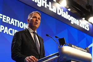 A man in a black suit with a blue tie speaking at a podium in front of a blue background