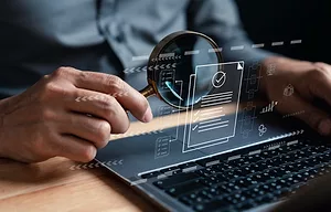 A PERSON HOLDS A MAGNIFYING GLASS IN FRONT OF AN OPEN LAPTOP. HOVERING ABOVE THE LAPTOP KEYBOARD IS A GRAPHIC OF A VIRTUAL PIECE OF PAPER WITH A CHECKMARK ON IT.