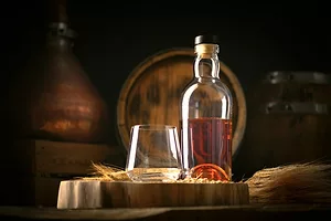 A clear bottle filled halfway with whisky, sitting on a barrel next to a rocks glass in front of a stack of other brown barrels