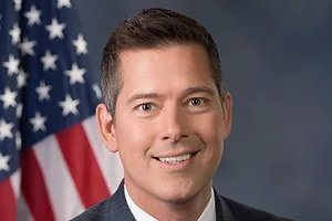 A MIDDLE-AGED WHITE MAN IN A SUIT AND TIE SMILES IN FRONT OF AN AMERICAN FLAG