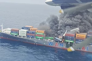 A navy blue container ship stacked with multi-colored containers, with thick black smoke billowing from the center section