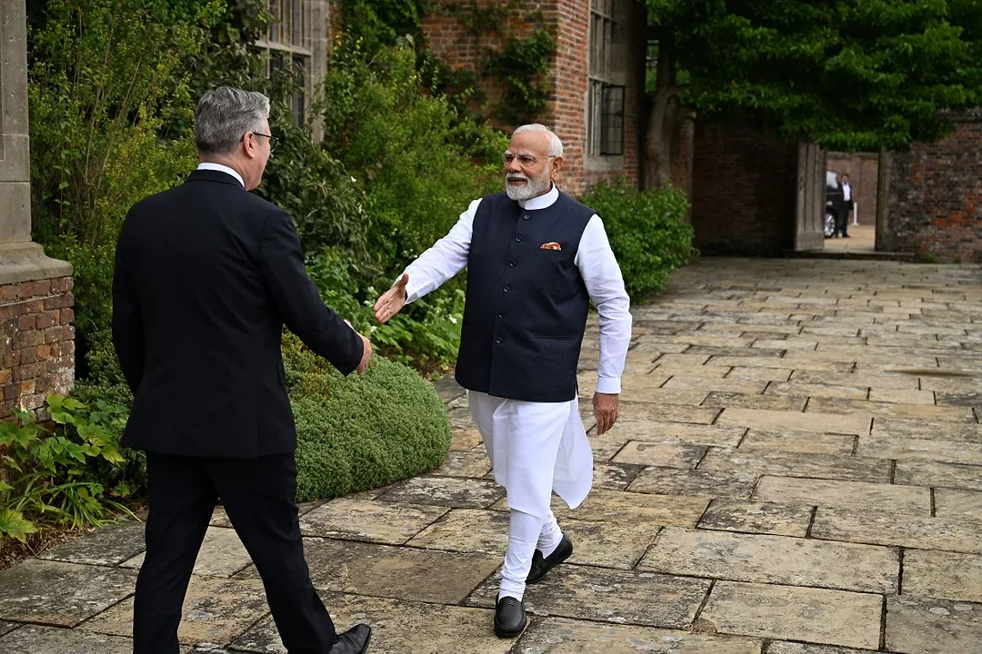 A MAN IN INDIAN CLOTHES WALKS TOWARDS A MAN IN A SUIT, HAND HELD OUT IN GREETING