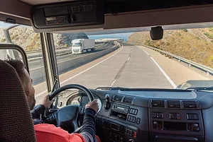 A PERSON IN A RED SHIRT IS DRIVING A TRUCK AND LOOKING OUT THE FRONT WINDOW OUT AT A HIGHWAY. ANOTHER TRUCK IS PASSING BY THE TRUCKER ON THE OTHER SIDE OF THE HIGHWAY.