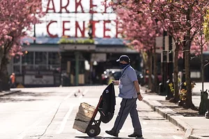A postal worker in a light blue shirt and navy blue cap, pushing a hand-cart stacked with white boxes across a city street