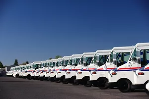 A FLEET OF UNITED STATES POSTAL SERVICE TRUCKS ARE PARKED NEXT TO EACH OTHER IN A PARKING LOT BELOW A CLEAR BLUE SKY.
