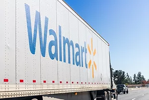 A LARGE WHITE WALMART TRACTOR TRAILER IS DRIVING ON A FREEWAY BEHIND A PICK-UP TRUCK ONE LANE OVER.