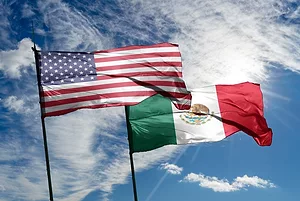 AMERICAN AND MEXICAN FLAGS FLUTTER AGAINST A CLOUD-STREAKED BLUE SKY